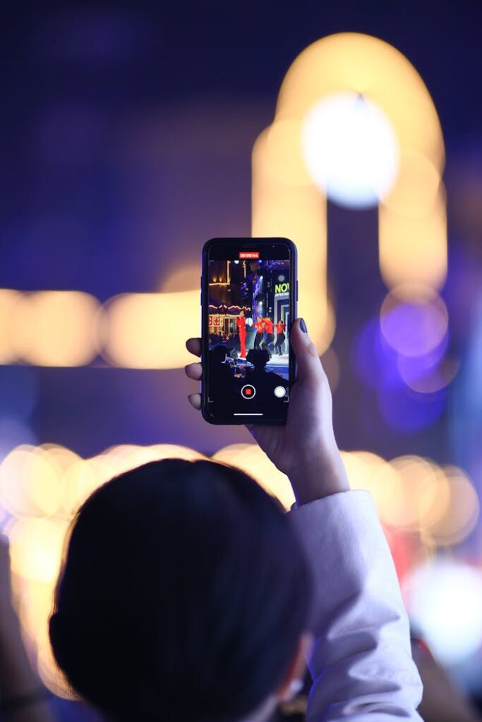 Person filming a vibrant night concert scene with smartphone, blurred festive lights in background.